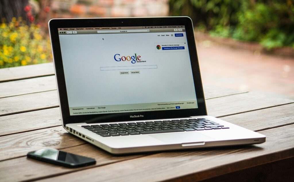 A MacBook Pro displaying Google Search engine on a wooden table outdoors, next to a smartphone.